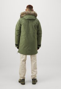 Parka verde oliva con cappuccio foderato in pelliccia, silhouette lunga, tasche con zip, polsini regolabili e una texture liscia. Indossata con pantaloni chiari e stivaletti.