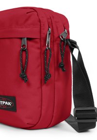 Röd tygaxelväska med två zippered fack, svarta remmar och silverdetaljer. Har en märkeslogopatch.