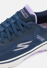 Navy blue Skechers Go Walk slip-on shoe with fabric upper, textured detailing, purple accents, and a white sole featuring rounded support nodes.