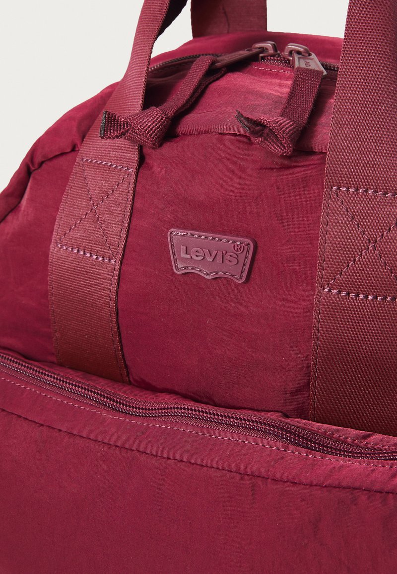 Maroone rygsæk med holdbart stof, der har et fremtrædende Levi's-logo, forstærkede håndtag og et lynlåslomme langs bunden.