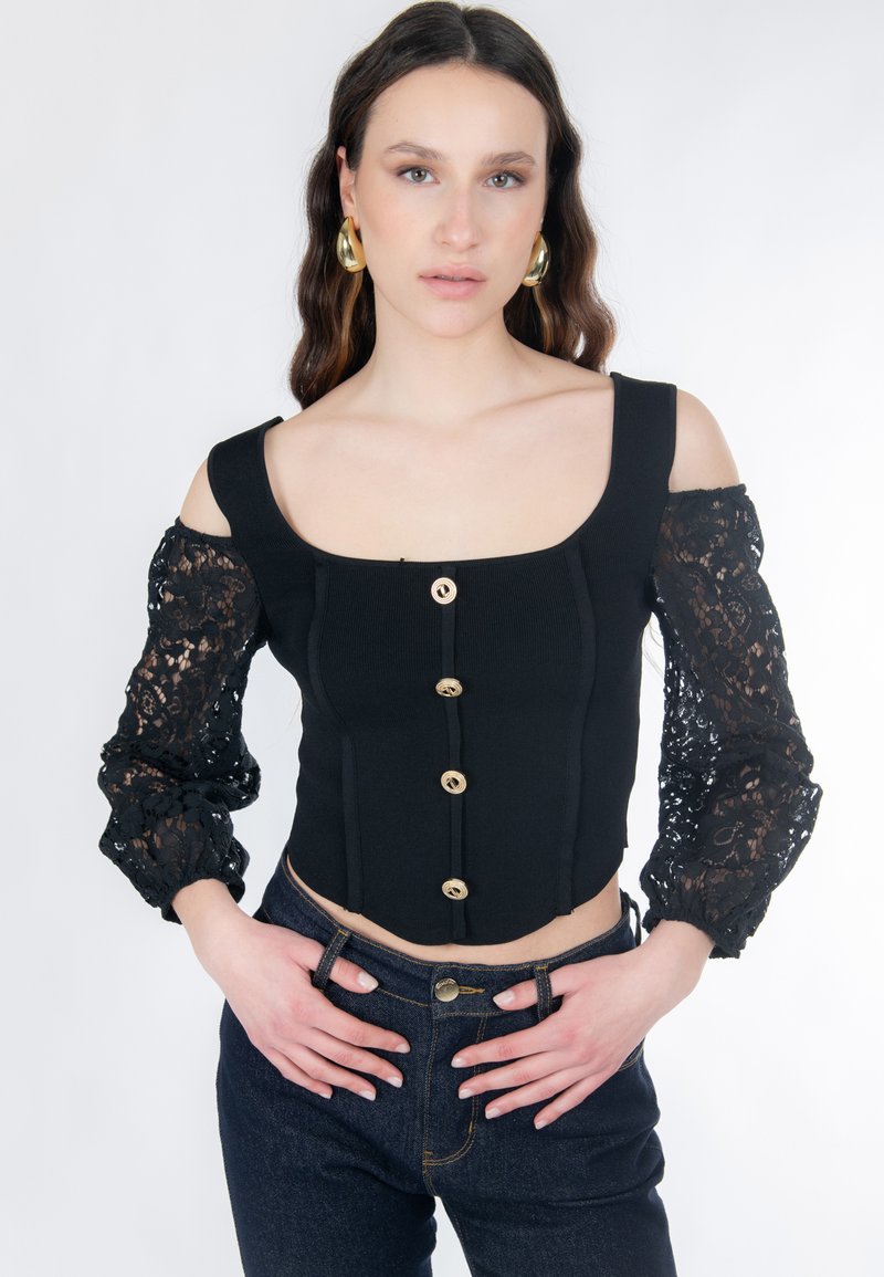 Schwarzes, kurzes Oberteil mit geripptem Körper und transparenten Spitzen-Volantsärmeln. Goldene Knöpfe akzentuieren die Vorderseite. Kombiniert mit dunkler Jeans vor neutralem Hintergrund.