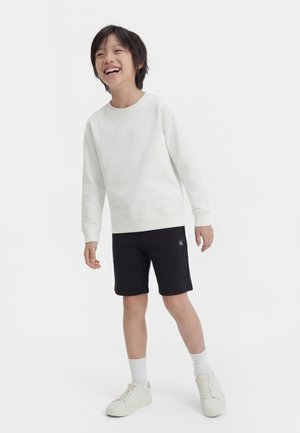 Usmievajúce sa dieťa stojí, oblečené v bielej košeli s dlhým rukávom, čiernych šortkách s logom, bielych ponožkách a bielych teniskách na jednoduchom pozadí.