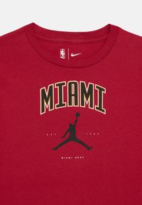 Sarkans kokvilnas T-krekls ar uzrakstu "MIAMI" dzeltenā un melnā krāsā, zem kura atrodas melns Jordana siluets un teksts "MIAMI HEAT". Iekļauti NBA un Nike logo.