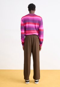 Flerfärgad stickad tröja med horisontella ränder i rosa och lila, ihop med bruna texturerade byxor. Skorna har en vit sula.