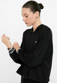 Čierna mikina s kapucňou vyrobená z mäkkej látky, s voľným strihom, zlatými doplnkami, šnúrkou na kapucni a kontrastnými vzorovanými manžetami.