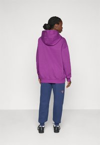 Un sweat à capuche violet avec une coupe décontractée, associé à un jogging bleu marine comportant un logo orange, porté avec des chaussures de sport noires.