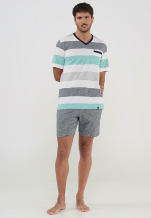 Hombre descalzo de pie, lleva una camiseta de rayas con cuello en V de manga corta y pantalones cortos grises, blancos y teal, frente a un fondo claro y liso.