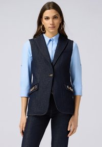 Dunkelblaues Denim-Weste mit Schalkragen, zwei Fronttaschen und goldfarbenen Akzenten. Über einem hellblauen Hemd mit Kragen getragen.