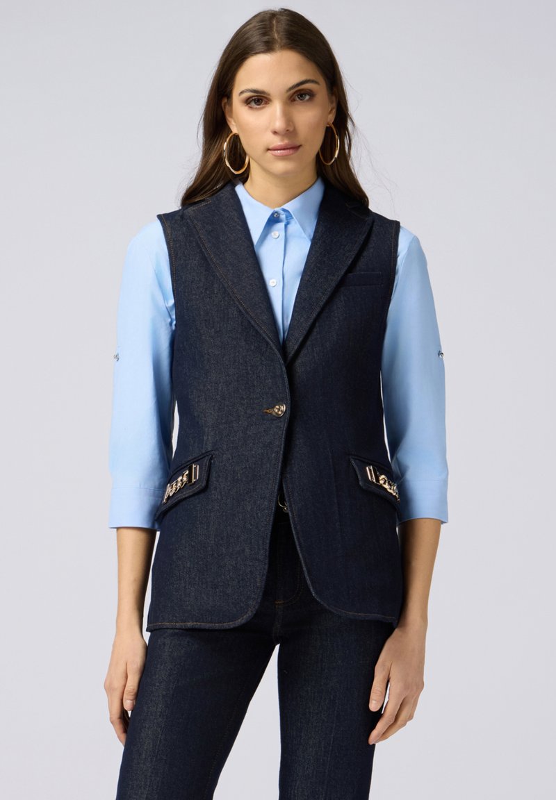 Dunkelblaues Denim-Weste mit Schalkragen, zwei Fronttaschen und goldfarbenen Akzenten. Über einem hellblauen Hemd mit Kragen getragen.