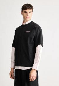 T-shirt preta de algodão com mangas curtas e gola redonda, apresentando um logótipo subtil em rosa no lado esquerdo do peito. Usada por cima de uma camisa rosa clara.