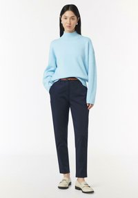 Hellblaues Turtleneck-Sweatshirt, lockere Passform; marineblaue Anzughose; brauer Gürtel; beige Slip-On-Schuhe mit schwarzen Sohlen. Minimalistisches Design, strukturierter Strick.