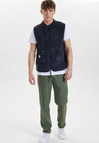 Marinblå fleeceväst med hög krage, zip-fickor på sidorna, bärs över en vit t-shirt, ihopmatchad med olivgröna byxor och vita sneakers.