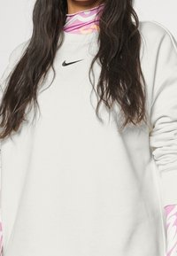 Sudadera blanca de manga larga, con un logotipo negro de Nike en el centro. Un cuello alto de color rosa con diseño es parcialmente visible debajo.
