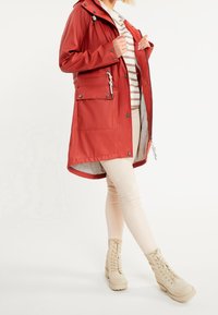 Red waterproof jacket with a hood, zip front, two front pockets, and snap closures. Worn over a striped shirt and beige pants with beige boots.