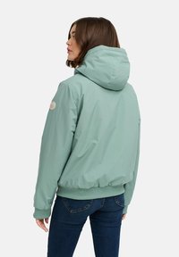 Gai zaļš kapuci jaka ar gludu tekstūru, elastīgu apkakli un apakšmalu, kā arī ar apzīmējumu uz kreisā pleca.