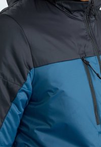 Vadderad jacka med en svart övre del och en blå nedre del. Har en hel dragkedja fram och en texturerad blå tyg.