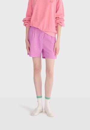 Person iført en lyserød sweatshirt, lilla shorts, hvide sokker med grønne striber og gennemsigtige slip-on sko, stående mod en ensfarvet baggrund.