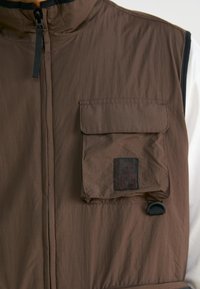 Brown insulated vest featuring a zip closure, high collar, front flap pocket with logo, and lightweight fabric texture.