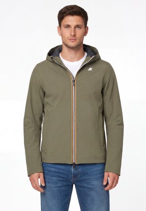 Mężczyzna w oliwkowo-zielonej kurtce z kapturem i zamkiem błyskawicznym z przodu, noszący białą koszulę i niebieskie dżinsy, stojący na białym tle.