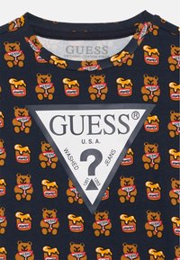 Tamsiai mėlynas medvilninis marškinėliai su pakartotu lokio ir medaus stiklainio raštu. Apdaila trikampiu baltų logotipų pleistru su tekstu "GUESS U.S.A.".