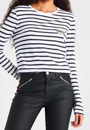 Femme portant un crop top à manches longues blanc et bleu marine rayé avec un patch étoile jaune et un pantalon noir taille haute avec des poches zippées.