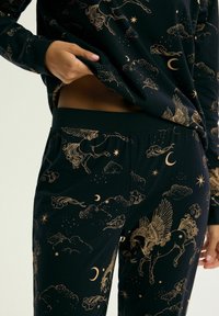 Ręce unoszą ciemny materiał piżamy ozdobiony złotym pegazem, gwiazdami, półksiężycami i wirującymi chmurami na bluzie i spodniach.