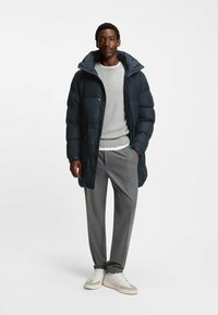 Giubbotto imbottito blu navy con cappuccio, maglione grigio, pantaloni grigi a fantasia e sneaker bianche. Dettagli notevoli su bottoni e cerniere.