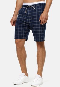 Pantalones cortos de cuadros marinos hechos de una tela suave, con una cinturilla elástica con cordón, patrón de rejilla blanca y bolsillos laterales. Combinados con zapatillas blancas.