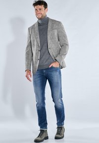 Grauer karierten Blazer über einem dunklen Rollkragenpullover, kombiniert mit hellen Jeans und grünen Wanderschuhen. Glatte Texturen mit figurbetontem Design.