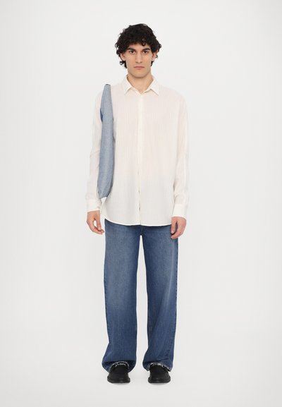 Camisa a rayas blancas, pantalones de mezclilla azules holgados y zapatos negros. La camisa tiene un diseño de botones y el conjunto presenta un aspecto casual.