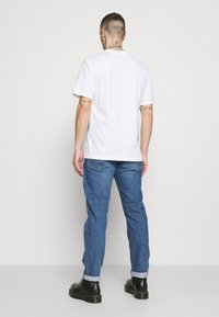 Vit kortärmad T-shirt med avslappnad passform, i kombination med ljusblå denimjeans och svarta ankelskor, sedd bakifrån.