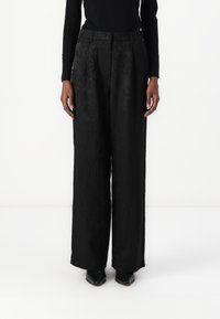 Pantalons large noirs en tissu à motifs, présentant une taille haute et des plis à l'avant. Associés à des chaussures noires, les mains reposant sur les côtés.