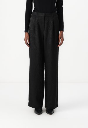 Zwarte wijde broek van gestructureerde stof, met een hoge taille en voorplooien. Gepaard met zwarte schoenen, met de handen aan de zijkanten.