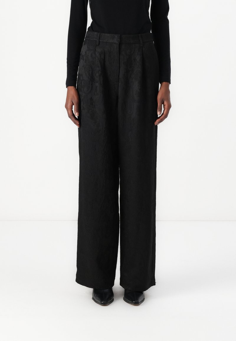 Pantalons large noirs en tissu à motifs, présentant une taille haute et des plis à l'avant. Associés à des chaussures noires, les mains reposant sur les côtés.