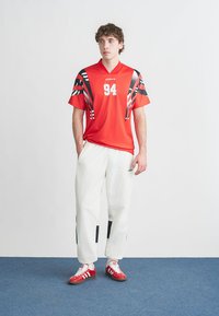 Röd atletisk tröja med svarta och vita detaljer och nummer 94; kombinerad med vita byxor med svarta detaljeringar; röda sneakers.