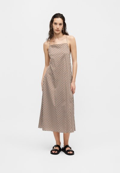 Femme aux cheveux mouillés portant une robe sans manches, longue jusqu'aux chevilles, à motif géométrique et des sandales noires, debout sur un fond blanc uni.