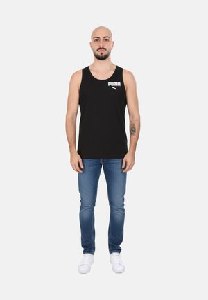 Uomo con barba e tatuaggio che indossa una canottiera nera Puma, jeans blu e sneakers bianche, in piedi davanti a uno sfondo bianco.