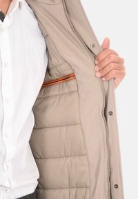 Giacca beige con chiusura a zip e bottoni a pressione, caratterizzata da un interno trapuntato, dettaglio della zip in accento arancione e una superficie esterna liscia.