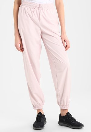 Hellrosa Jogginghose mit elastischem Bund und Bündchen, getragen mit schwarzen Sportschuhen an einer stehenden Person vor weißem Hintergrund.