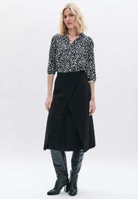 Gonna nera avvolgente abbinata a una blusa a fantasia nera e crema. Il modello indossa stivali neri in pelle al ginocchio. Accenti di gioielleria semplici.
