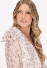 Blouse in lichtgewicht, transparant materiaal met pastel bloemenpatroon. Bevat rimpelende schouderdetails en een V-hals. Zachte, vloeiende textuur.