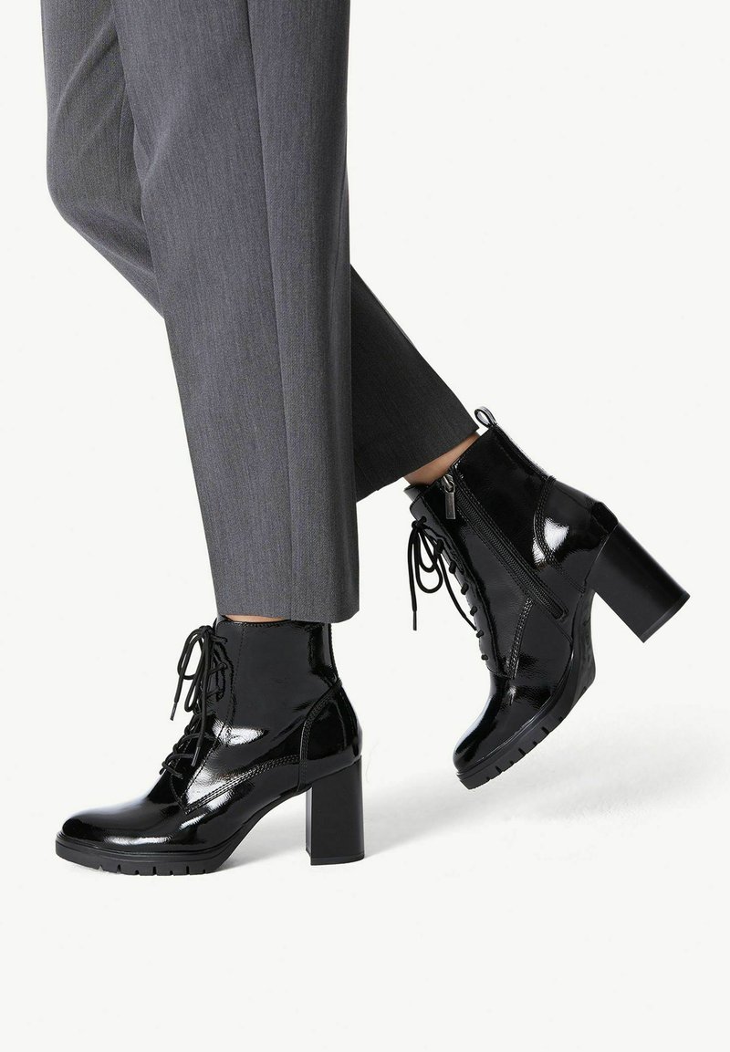 Bottines noires en cuir verni avec un talon épais et un devant à lacets. Comprend une fermeture éclair sur le côté et une semelle texturée. Associées à un pantalon gris ajusté.
