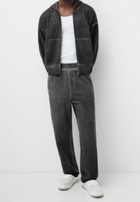 Sudadera con capucha de velour gris con bolsillos delanteros, combinada con pantalones deportivos grises a juego. La modelo lleva una camiseta sin mangas blanca y zapatillas ligeras. Diseño sencillo.