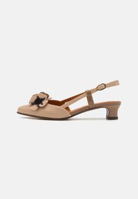 Chaussure à bride slingback en cuir beige avec un bout pointu, un petit talon carré, et un accent floral décoratif sur le bout. La chaussure comprend une sangle de cheville ajustable.