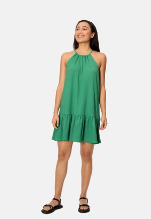 Mujer de pie sonriendo, vestida con un vestido verde sin mangas tipo halter con volante en el dobladillo y sandalias negras de tiras, sobre un fondo liso.