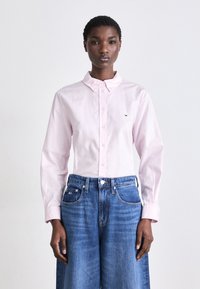 Camicia button-up rosa chiaro a maniche lunghe con sottili righe, abbinata a jeans a gamba larga in denim blu a vita alta. Presenta un piccolo logo sulla camicia.