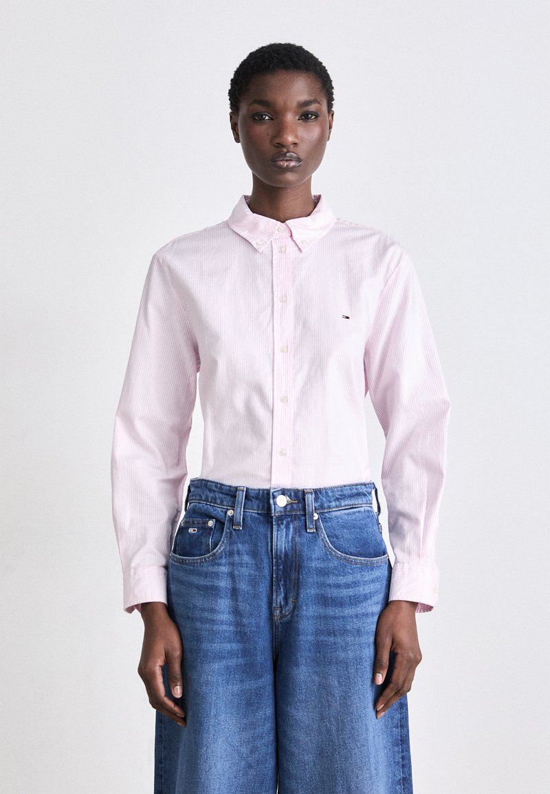 Camicia button-up rosa chiaro a maniche lunghe con sottili righe, abbinata a jeans a gamba larga in denim blu a vita alta. Presenta un piccolo logo sulla camicia.