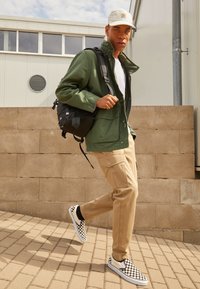 Veste en toile verte, T-shirt blanc, pantalons cargo beiges, sac à dos noir, et chaussures à enfiler à carreaux noir et blanc. Marchant sur un chemin carrelé.