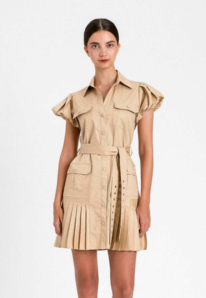 Donna che indossa un vestito camicia beige a maniche corte con cintura, gonna plissettata e tasche con patta, in piedi su uno sfondo bianco semplice.