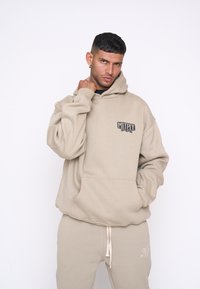 Felpa beige con tasca frontale e patch con logo. Il tessuto sembra morbido e ha una vestibilità ampia. Abbinata a pantaloni beige coordinati.